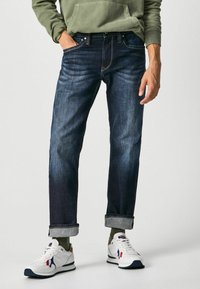 Jeans de mezclilla azul oscuro con un corte relajado, con puños enrollados. Combinados con zapatillas blancas y una sudadera verde oliva para un look informal.