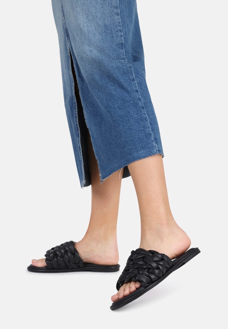 Sandales à enfiler noires tissées avec semelle plate et design à bout ouvert. Associées à une jupe en denim bleu avec une fente latérale et un ourlet brut.