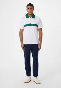 Poloshirt i hvid bomuld med grøn krave, der har en central stribe i grøn og rød. Sammen med marineblå bukser og hvide sneakers.