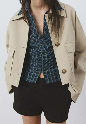 Mujer con chaqueta beige abotonada sobre blusa abullonada de cuadros azul verdosos y pantalones cortos negros, con las manos en los bolsillos de la chaqueta, sobre un fondo liso.