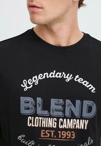 Juodas medvilnės marškinėliai su grafiniu tekstu balta, mėlyna ir oranžine spalvomis. Apvalus kaklas. Tekstas apima "LEGENDARY TEAM", "BLEND" ir "EST. 1993."