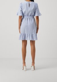 Robe à rayures bleues et blanches avec des manches courtes à volants, un dos échancré en forme de goutte et un ourlet évasé. Portée avec des sandales blanches à talons blocs.