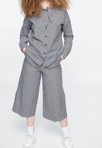 Musta-valkoinen ruudullinen, napitettava paita, jossa kaulus, yhdistetty samanlaisiin leveälahkeisiin, lyhennettyihin housuihin. Malli käyttää valkoisia tennareita.