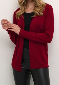 Rode cardigan met lange mouwen, open voorkant ontwerp, zachte textuur en lichte rek; gedragen over een zwart topje en gecombineerd met zwarte broek.