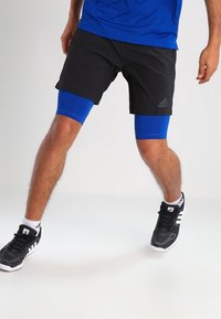 Svarta träningsshorts med blå kompressions leggings underneath, bärs med svart och vita sportskor. Har Adidas-logotyp på shortsen.