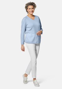Helles blaues V-Ausschnitt-Pullover mit langen Ärmeln, kombiniert mit weißen, schmal geschnittenen Hosen und grauen Turnschuhen. Glatte Struktur mit Seitennahtdetails.