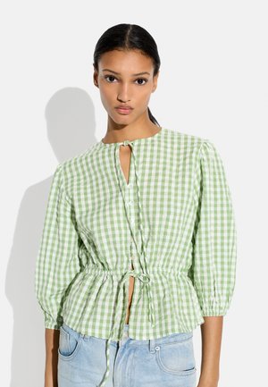 Młoda kobieta ubrana w jasnozieloną i białą koszulę w kratę z guzikami i wiązaniami z przodu, połączoną z jasnoniebieskimi dżinsami.