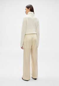 Γυναίκα με μακριά σκούρα μαλλιά, που φοράει λευκό πουλόβερ με ζιβάγκο και άνετο μπεζ παντελόνι, στεκόμενη με την πλάτη προς τα εμπρός σε λευκό φόντο.