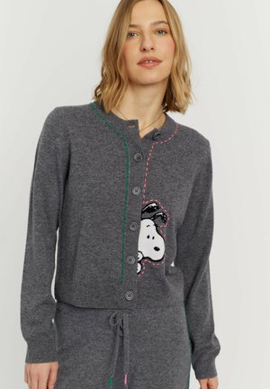 Vrouw draagt een grijze vest met knopen met Snoopy-cartoon en bijpassende broek met trekkoord op een lichte achtergrond.