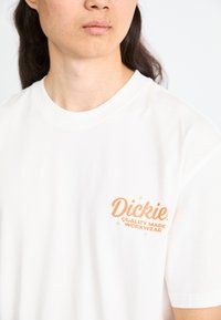 Witte katoenen T-shirt met korte mouwen en een ronde halslijn. Ontwerp aan de voorkant bevat oranje en groene tekst met een logo.