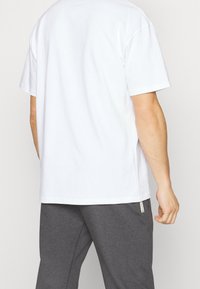 Biały bawełniany t-shirt o luźnym kroju, z krótkimi rękawami i zaokrąglonym dołem, w zestawie z szarymi dresami z zamkami błyskawicznymi w kieszeniach.