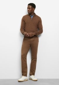 Brauner Pullover mit Reißverschluss am Kragen und gerippten Bündchen, kombiniert mit braunen Chinos und weißen Sneakern. Einfaches Design mit lockerer Passform und neutralen Farbtönen.
