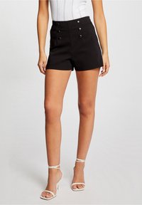 Shorts noirs taille haute avec une coupe ajustée, dotés de doubles boutons à l'avant et d'une texture de tissu lisse. Associés à des talons blancs à brides.