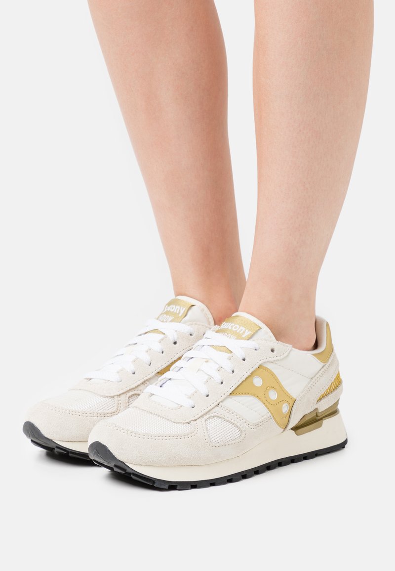 Beige en witte sportchoenen met suède en mesh materiaal, gouden accenten, veters, geperforeerde details en een zwarte rubberen zool.
