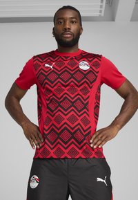 Rdeča športna majica s črnim geometrijskim vzorcem, kratkimi rokavi, logotip Puma na prsih in ramu, kombinirana z črnimi kratkimi hlačami z logotipom.