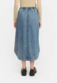 Denim midi krilo z elastičnim pasom, modre barve, gladke teksture in širokim robom. Črna obutev dopolnjuje obleko.