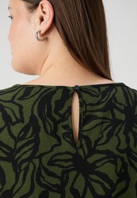 Grön mönstrad topp med ett svart bladdesign, med en nyckelhålsöppning bak som hålls på plats med en knapp. Texturerad tyg.