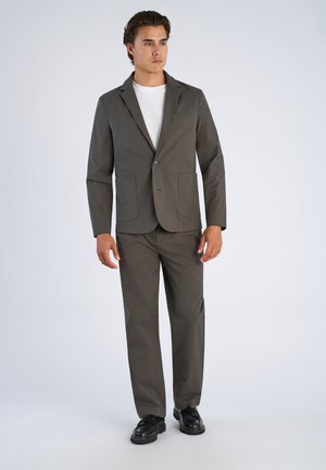 Jonge man die een donkergrijs pak met twee voorzakken, een wit overhemd en zwarte schoenen draagt, staand tegen een effen lichte achtergrond.