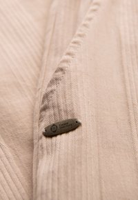 Tissu beige texturé avec des rayures verticales, muni d'une petite étiquette métallique portant la mention "OMBRE HERITAGE" attachée au matériau.