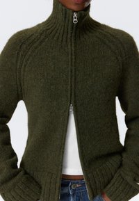 Cárdigan de punto verde con cuello alto, cierre de cremallera en la parte delantera, puños acanalados y dobladillo. Textura suave con un ajuste ligeramente holgado.