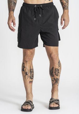 Homme portant un short cargo noir avec taille à cordon, plusieurs tatouages sur les jambes et les bras, et des sandales grises à enfiler avec un texte noir.