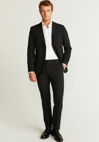 Sort jakkesæt med notchtaljer, parret med en hvid skjorte. Modellen står selvsikkert iført sorte dress sko.
