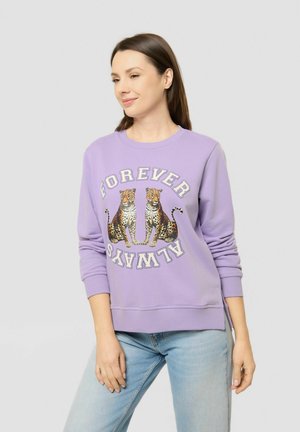 Woman wearing a light purple sweatshirt with two leopards and the text "FOREVER ALWAYS" paired with light blue jeans against a plain background.