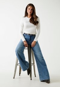 Witte lange mouw shirt met grafische print, gecombineerd met lichtblauwe wijd-uitlopende jeans en donkere haklaarzen, zittend op een metalen kruk.