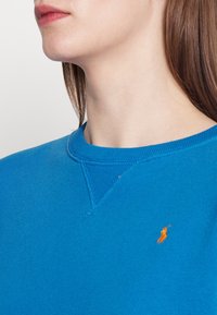 Blå sweatshirt i bomullsblandning med rund halsringning, ribbad krage och en orange broderad logotyp på bröstet. Slät textur.