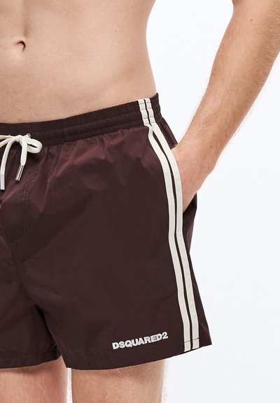 Bañador marrón con cintura elástica, cordón blanco y dos franjas laterales blancas. Presenta un logo "DSQUARED2" en el dobladillo.