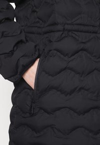Zwarte gewatteerde jas met een ritzak en een gathered elastische tailleband. De stof heeft een gladde textuur met eengequilt patroon.