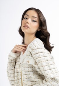 Cremefarbener Cardigan mit strukturiertem Gewebe, goldenen Akzenten, einem V-Ausschnitt und einem dekorativen Perlenknopf an der Tasche.