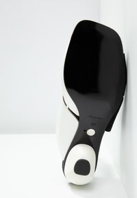 Semelle d'une chaussure à talon noire et blanche pour femme, avec un bout carré, un dessous noir lisse et un talon blanc avec une pièce noire.