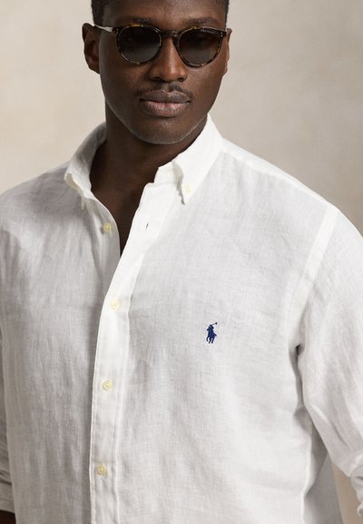 Mężczyzna noszący okulary przeciwsłoneczne w oprawie w deseń żółwia oraz białą koszulę na guziki z małym niebieskim logiem polo na piersi.