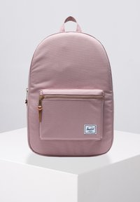 Mochila rosa claro hecha de tejido duradero, con forma redondeada, un bolsillo frontal con cremallera y herrajes dorados, y un asa en la parte superior.