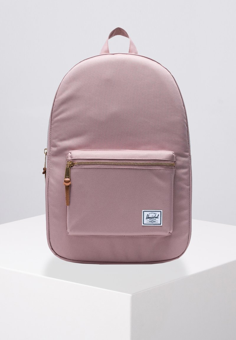 Mochila rosa claro hecha de tejido duradero, con forma redondeada, un bolsillo frontal con cremallera y herrajes dorados, y un asa en la parte superior.