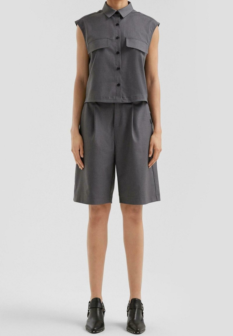 Personne portant une chemise grise sans manches avec poches sur la poitrine, un short assorti jusqu’aux genoux et des bottines noires, debout devant un fond uni.