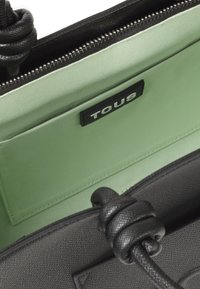 Czarna, teksturowana torebka z węzłowymi uchwytami, otwarta, by pokazać jasnozieloną podszewkę oraz wszytą kieszeń z czarną metką marki TOUS.