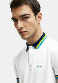 Polo bianco con colletto a righe blu navy, giallo e azzurro. Presenta un piccolo logo ricamato sul petto e dei bottoni.