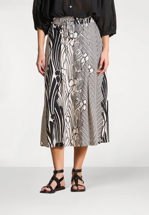 Femme portant une jupe midi à motifs noir et blanc avec une taille nouée, un chemisier noir et des sandales noires à brides, debout sur un sol gris.