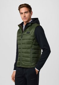 Zielona kamizelka puchowa z zapięciem na zamek, mająca stojący kołnierz, czarne akcenty i naszywkę z logo. Noszona na czarnej koszulce z długim rękawem.