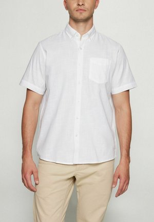 Mand iført en kortærmet hvid skjorte med brystlomme og beige bukser, stående foran en ensfarvet baggrund.