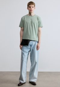 Light green cotton t-shirt with small logo, paired with loose light blue denim jeans and a black accent bag at the waist. Dark brown shoes.