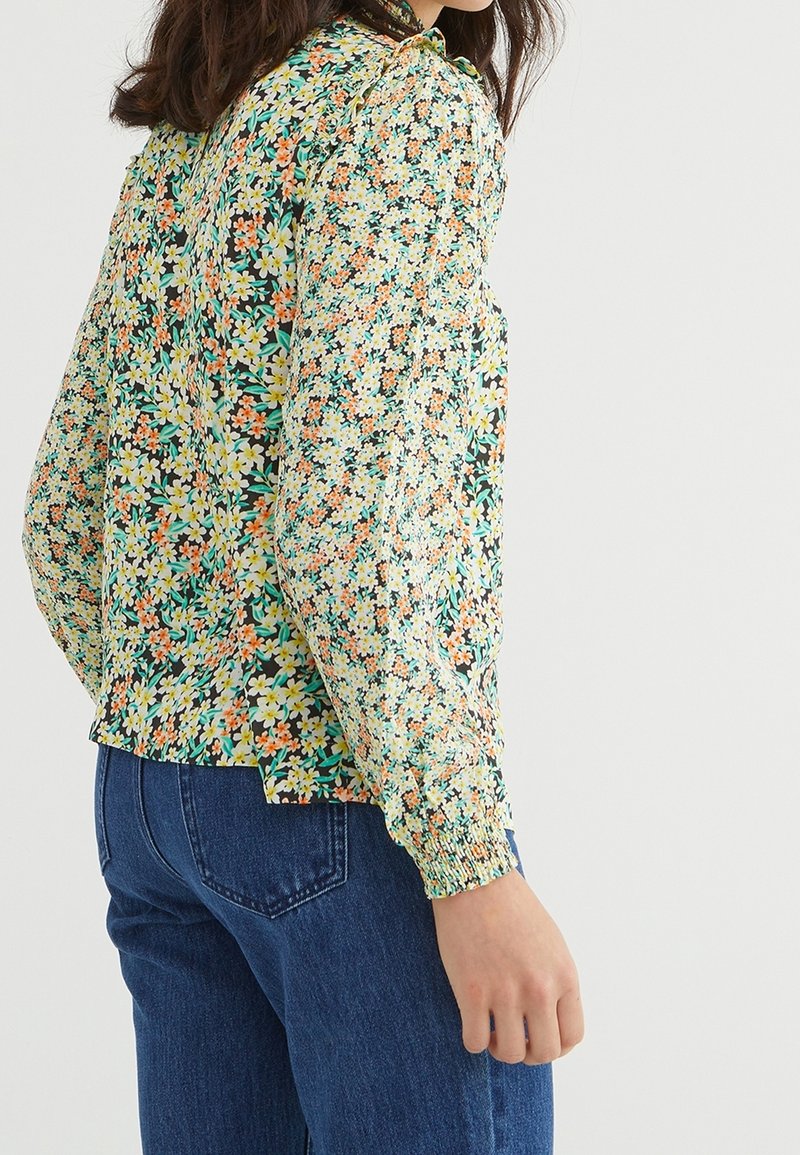 Blusa floreale verde, con un motivo di fiori multicolori, maniche lunghe con polsini arricciati e un collo leggermente arricciato.