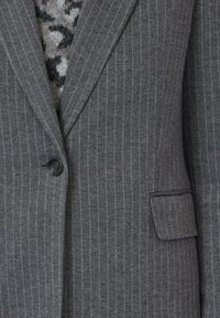 Grijs pinstripe blazer met een enkele zwarte knoop, een klepzak en een gebreide trui met patroon zichtbaar daaronder.