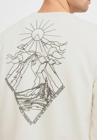Kermansweater med en linjetecknad design av berg, moln och sol på baksidan, med texten "Vandrade tankar - vilda tankar."