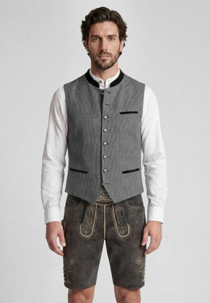 Mann, der traditionelle bayerische Tracht mit grauer Weste, weißem Hemd und dunkelbraunen bestickten Lederhosen trägt, steht vor einem schlichten Hintergrund.