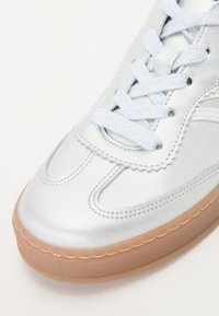 Stříbrná kožená sneakers s hladkým povrchem, kulatou špičkou, bílými šňůrkami a hnědou gumovou podešví s texturovaným okrajem.