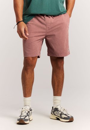Man draagt textuurrijke, stoffige roze short met trekkoord, groene blouse, witte enkelsokken en grijs-witte sportieve sneakers, staat met handen in de zakken.