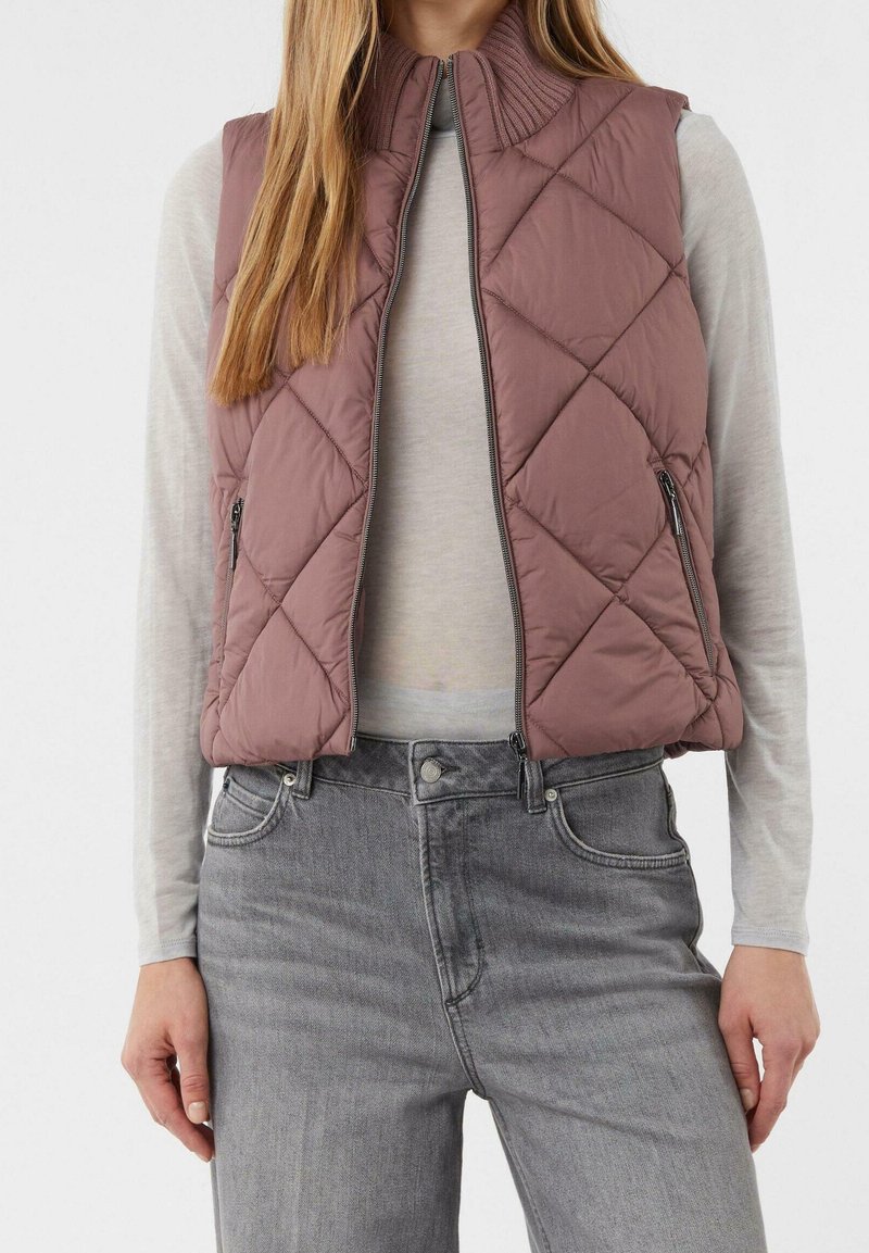 Femme portant un gilet matelassé mauve avec fermeture éclair, une chemise à manches longues gris clair et un jean gris taille haute, debout devant un fond uni.